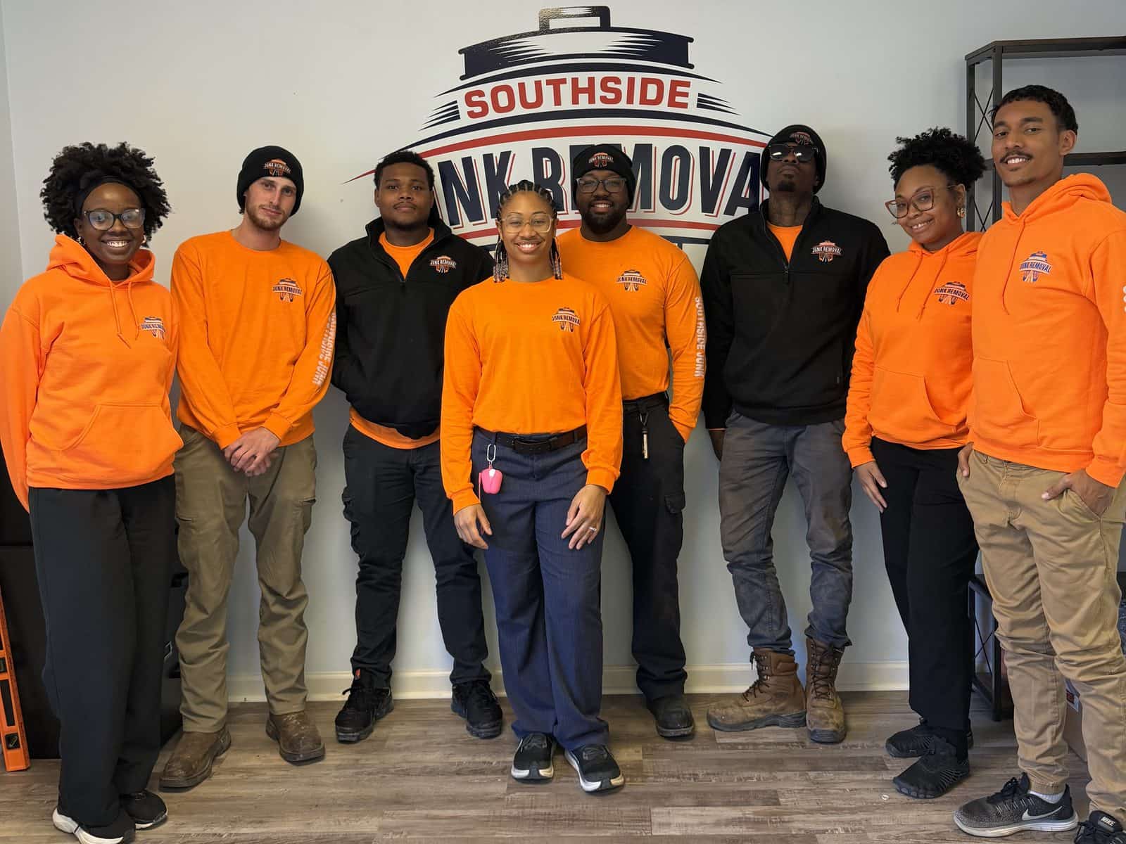 The Southside Junk Removal team in orange company shirts and hoodies in front of the logo wall at the Jonesboro office