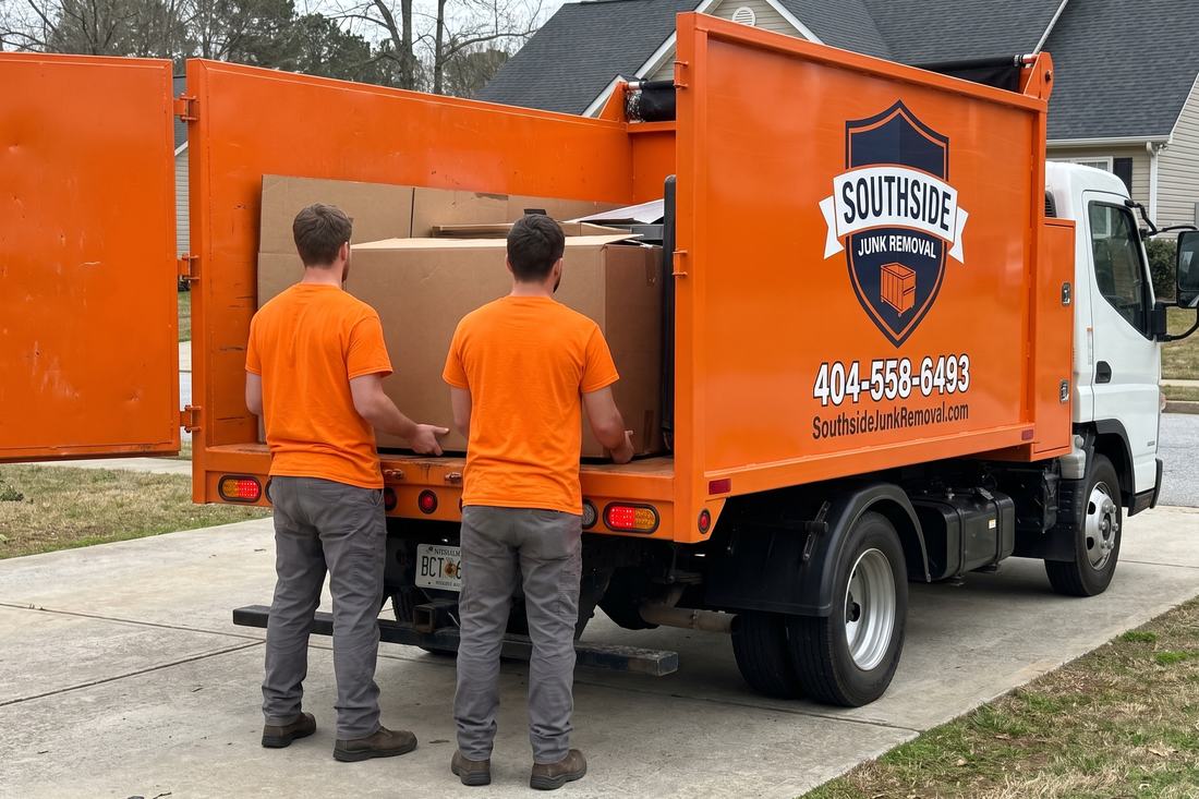 Two Southside Junk Removal crew members sliding a cardboard box through the open rear tailgate of the orange dump truck on a driveway at a Jonesboro, Georgia home