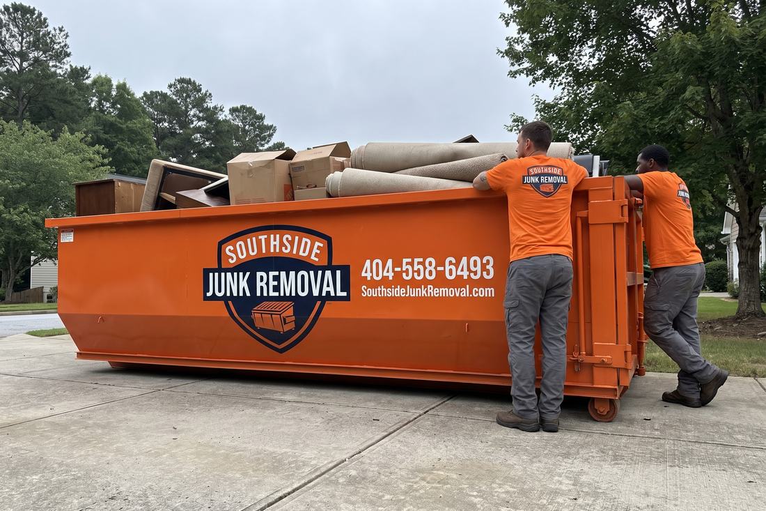 Two Southside Junk Removal crew members beside a half full orange roll-off dumpster holding furniture, cardboard boxes and rolled carpet on a Stockbridge, Georgia driveway